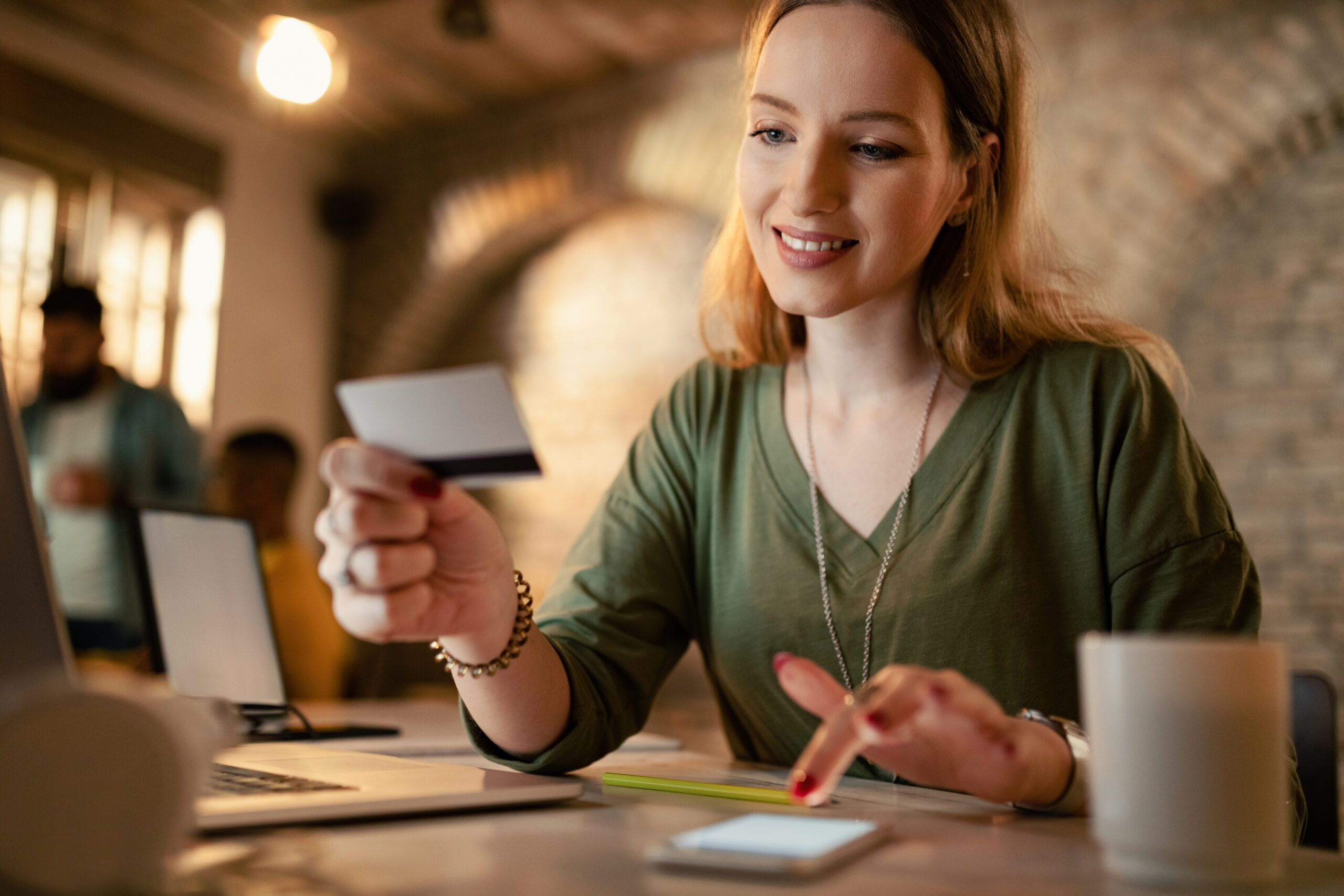 Uma mulher está sentada em uma mesa, sorrindo, segurando um cartão de crédito em uma das mãos e usando um smartphone com a outra. Um laptop e uma caneca de café estão sobre a mesa, e o plano de fundo mostra uma sala moderna e bem iluminada.