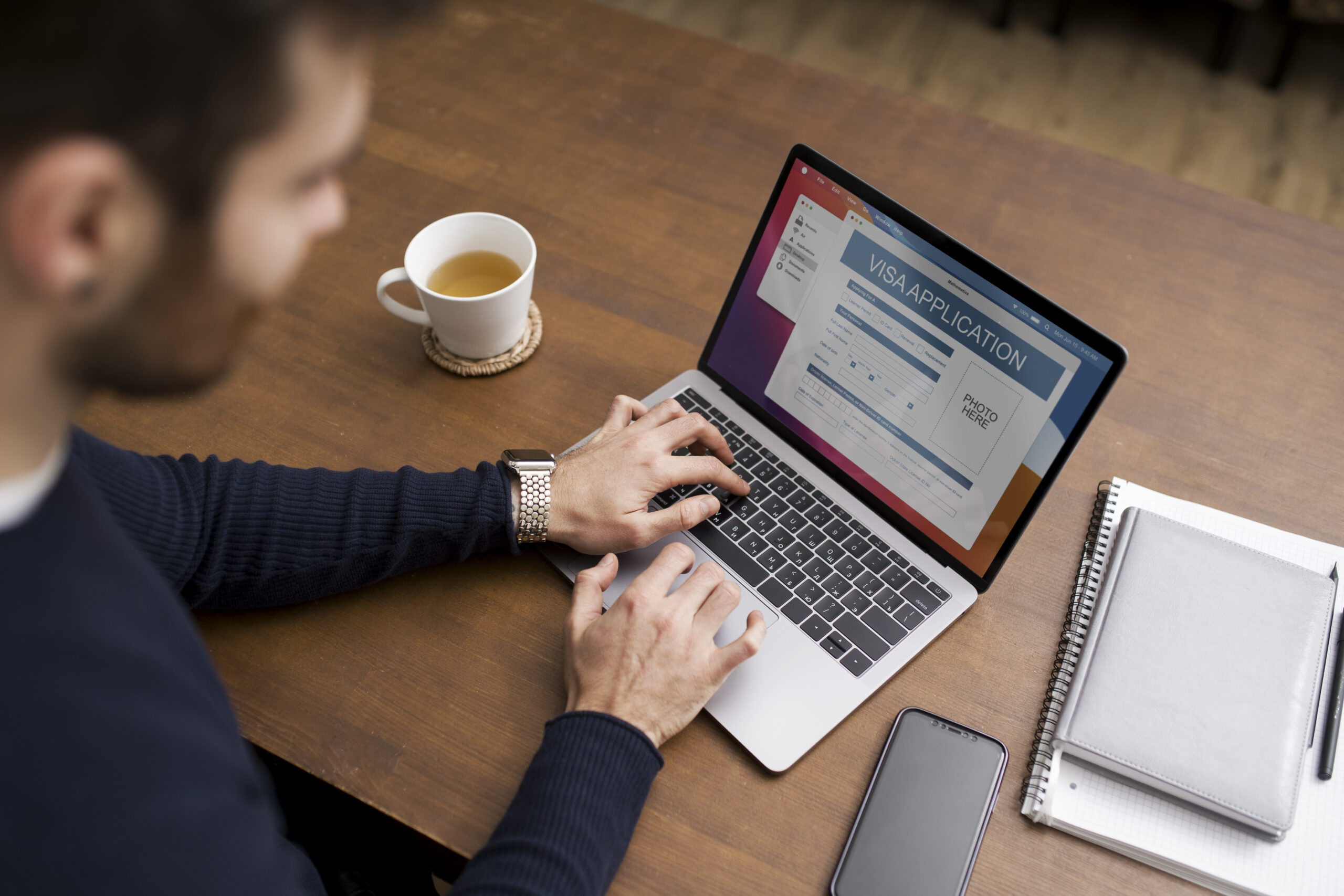 Uma pessoa digita em um laptop exibindo um formulário de solicitação de visto on-line. Uma xícara de chá, um smartphone, uma caneta e um caderno estão sobre a mesa de madeira ao lado.