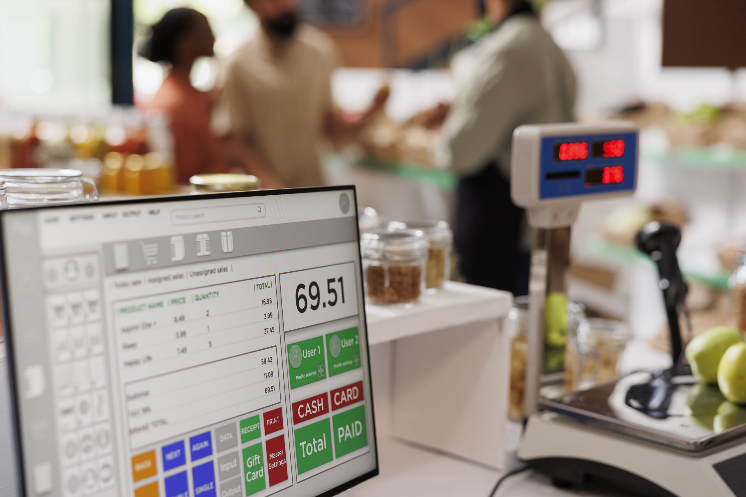 Um close-up de uma tela de caixa digital mostrando um total de 69,51 no balcão de um supermercado, com uma balança, maçãs e compradores desfocados ao fundo.