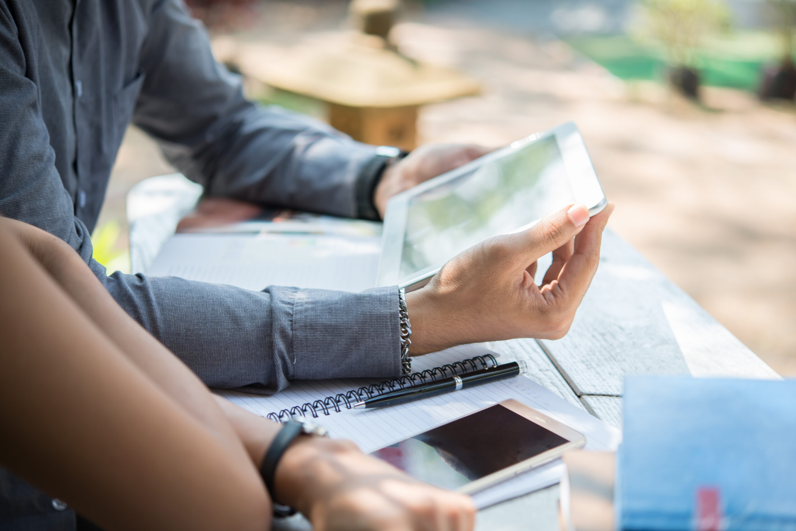 Duas pessoas sentadas em uma mesa ao ar livre com cadernos, um smartphone e um tablet, colaborando ou estudando. Uma pessoa segura o tablet enquanto a outra escreve em um caderno espiral.