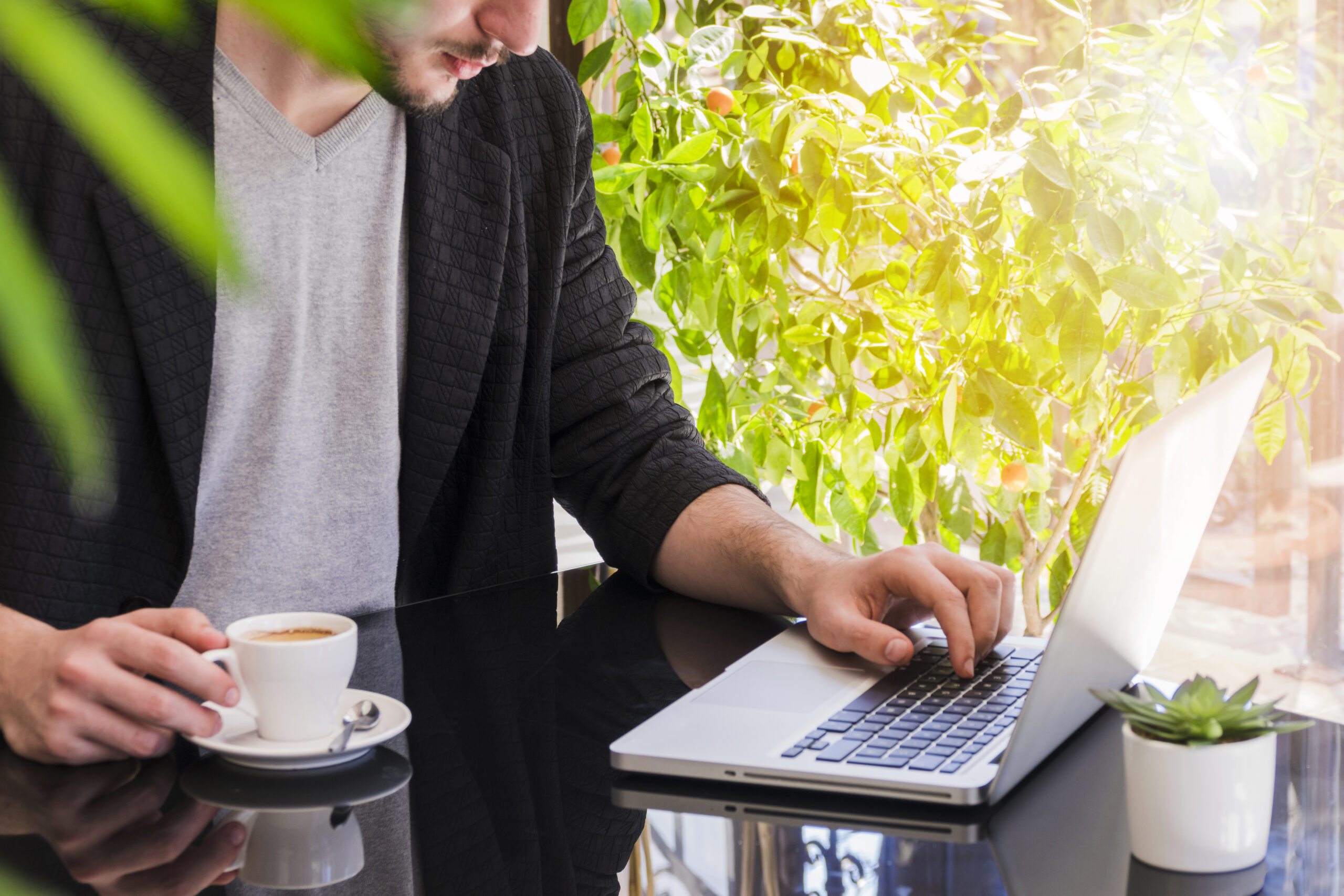 Uma pessoa está sentada em uma mesa preta reflexiva, usando um laptop com uma das mãos e segurando uma xícara de café com a outra. O ambiente é claro, com plantas verdes ao fundo e uma pequena planta sobre a mesa.