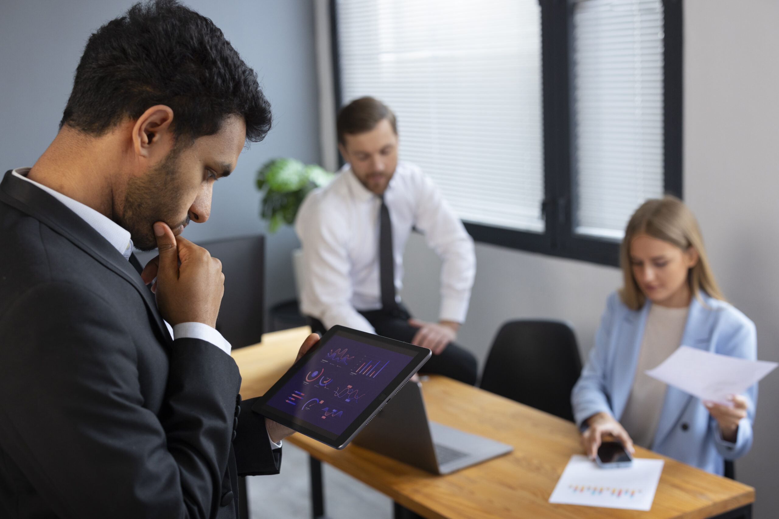 Um homem de terno estuda um tablet que exibe gráficos, enquanto um homem e uma mulher sentam-se em uma mesa atrás dele, olhando para documentos e um telefone em um ambiente de escritório moderno.