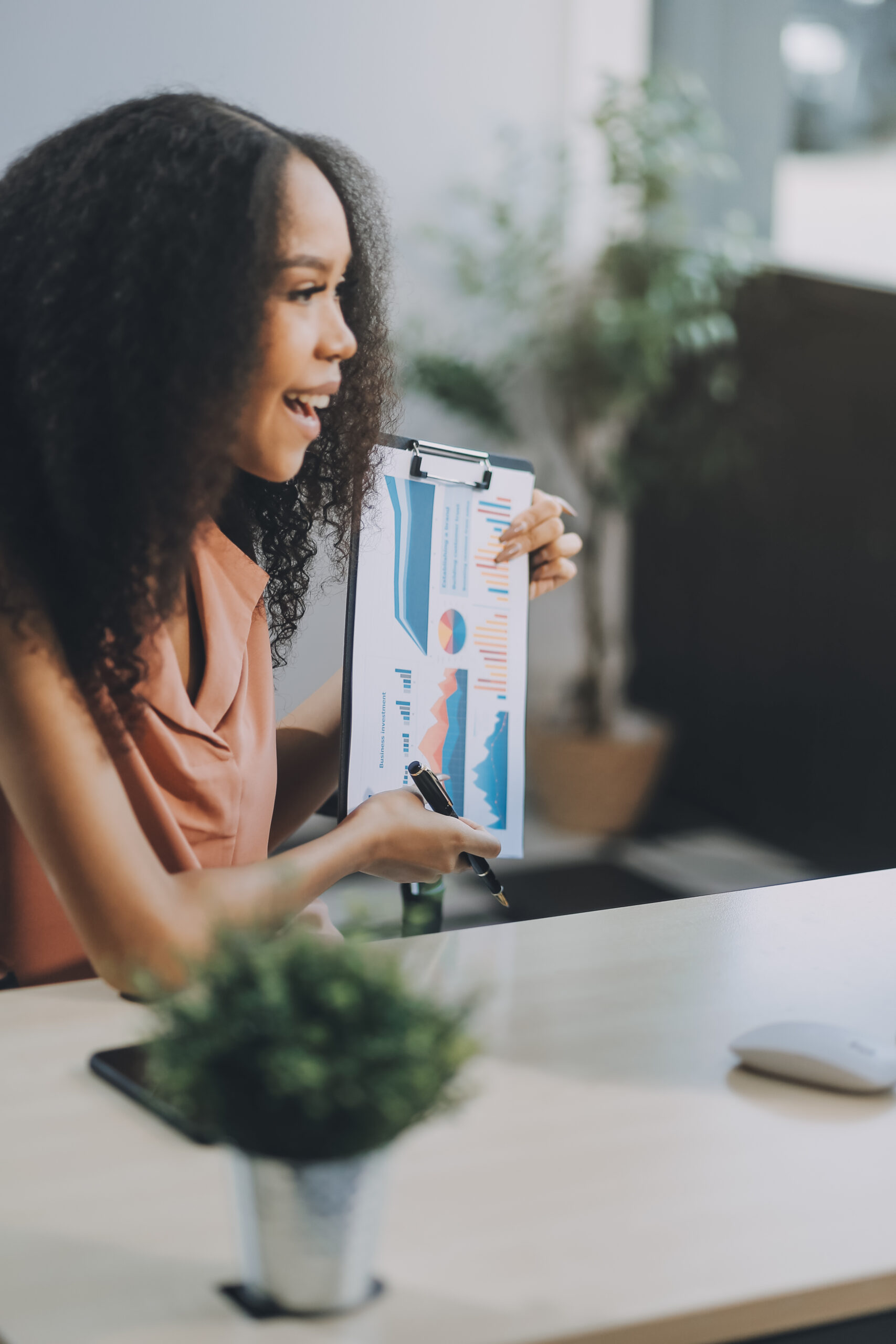 Uma mulher está sentada em uma mesa, sorrindo e segurando uma prancheta com tabelas e gráficos coloridos, gesticulando com uma caneta. Há plantas e materiais de escritório ao fundo.