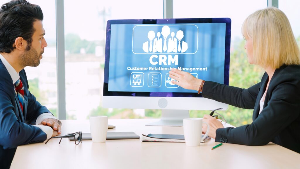 Dois profissionais de negócios sentados em uma mesa discutindo informações exibidas em uma tela de computador que mostra um gráfico de CRM Customer Relationship Management com ícones, em um cenário de grandes janelas.