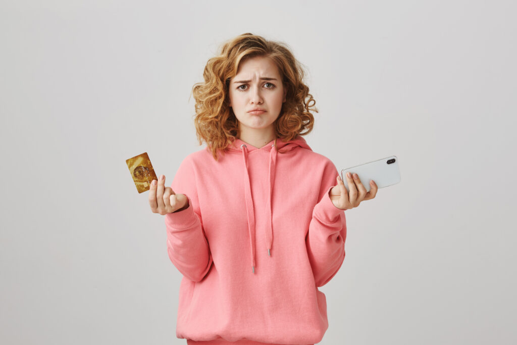 Uma jovem de cabelos cacheados com capuz rosa parece confusa, segurando um cartão de crédito em uma das mãos e um smartphone na outra, em um fundo claro e simples.
